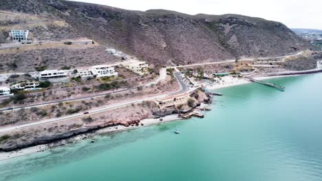 Panorama-aerial-view-of-Playa-El-Caymancito-nearby-coast-line-near-La-paz-Baja-California-Sur-Mexico-with-a-view-of-a-busy-road-right-on-the-sea,-rocky-landscape-with-buildings