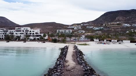 Luftaufnahme-Eines-Dollys-Vor-Der-Küste-Von-Playa-El-Caymancito-In-Der-Nähe-Von-La-Paz-Baja-California-Sur-Mexico-Am-Strand-Mit-Blick-Auf-Hotelgebäude,-Trockene-Landschaft-Und-Das-Blaue-Meer