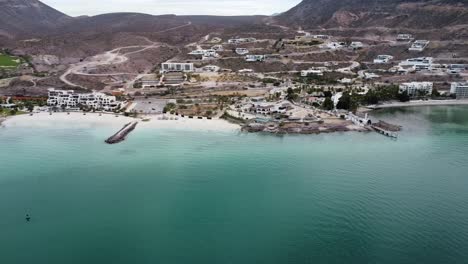 Aerial-panning-shot-over-the-beautiful-coast-of-Playa-El-Caymancito-near-La-paz-Baja-California-Sur-Mexico-with-view-of-dry-landscape-and-hotel-building-with-turquoise-sea
