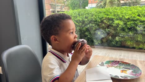 Cute-3-year-old-black-child-enjoys-eating-a-chocolate-muffin-in-a-cafeteria-next-to-the-window
