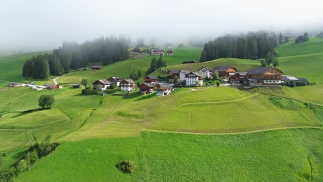 Nestled-amidst-verdant-fields,-the-houses-and-farms-in-Val-Badia-present-a-tranquil-portrait-of-rural-life,-with-a-soft-blanket-of-mist-gently-rolling-in-the-distance