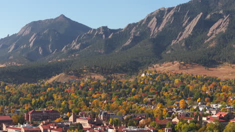 Aerial-up-close-view-of-Flatiron-mountains-in-Boulder-Colorado-surrounded-by-peak-fall-colors-of-green,-red,-and-yellow-trees-showing-the-town-and-CU-Boulder-campus