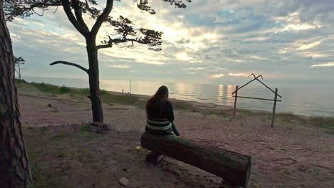 Lonely-woman-sitting-on-wooden-bench-on-sea-coastline,-back-moving-shot