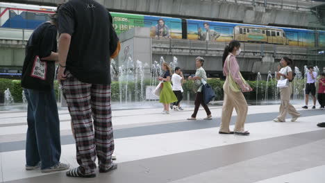 La-Gente-Está-Ocupada-Caminando-Fuera-De-Siam-Paragon-En-Bangkok-Con-La-Estación-De-Tren-BTS-Con-Trenes-Que-Salen-Y-Paran-Para-Pasajeros,-En-Tailandia