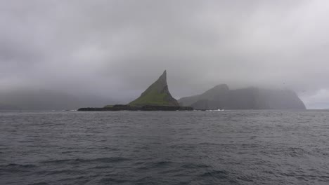 Boat-point-of-view-approaching-Tindholmur-on-moody,-cloudy-day,-Faroe-Islands