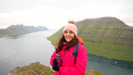 Woman-smiles-happily-while-admiring-view-from-Klakkur-Mountain-peak-holding-a-thermos,-Faroe-Islands
