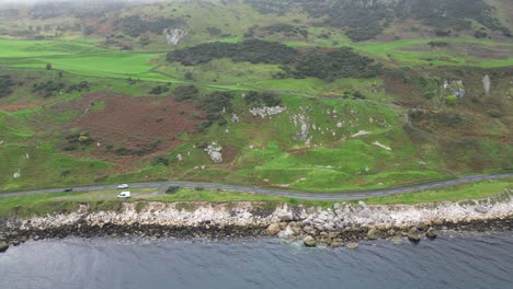 Aerial-panning-view-over-the-idyllic-rocky-coast-road-right-on-the-sea-with-moving-cars-during-an-adventurous-trip-near-glenarm-town-in-Northern-Ireland