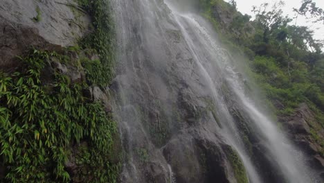 Tilt-down-misty,-wispy-waterfall-on-lush-green-jungle-rock-cliff