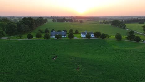 Ruhige-Pferdefarm-In-Kentucky-Bei-Sonnenuntergang:-üppige-Grüne-Felder,-Vereinzelte-Bäume,-Weiße-Bauernhäuser-Und-Ein-Entfernter-Orangefarbener-Horizont