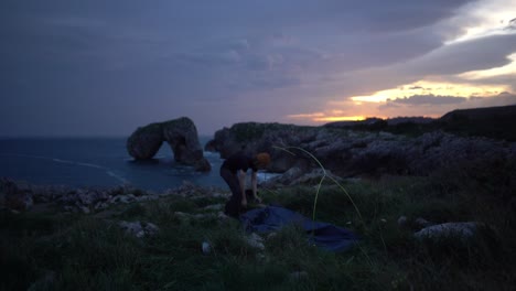 Young-adventurous-camping-man-packs-tent-at-sunrise-on-ocean-cliff