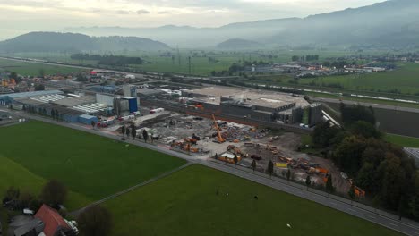 Aerial-view-showing-Construction-site-of-new-Läderach-Chocolate-factory-in-Bilten,-Switzerland---Foggy-fields-and-mountains-in-backdrop