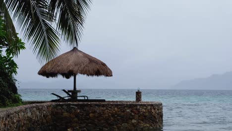 Lonely-thatched-roof-sun-parasol-with-nobody-around-on-a-remote-tropical-island-during-monsoon-season-with-grey,-wet-and-raining-weather-conditions
