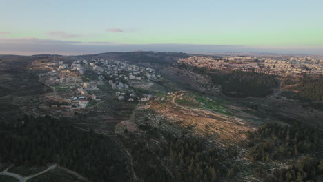 The-mountainous-area-of-Ramot-neighborhood-in-Jerusalem,-Israel-on-the-right-and-Beit-Iksa,-the-Palestinian-village-on-the-left-over-the-Kidron-Valley,-Israel---drone-sliding-shot
