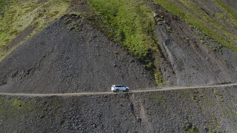 Toma-Aérea-De-Seguimiento-De-Una-Camioneta-Blanca-En-Una-Carretera-Peligrosa-En-El-Borde-De-Una-Montaña-Rocosa-En-Islandia.