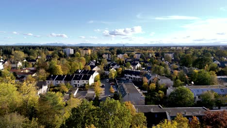 Aerial-view-over-Munich-suburbs:-Drone-glides-showcasing-crisp-autumn-forests-leading-to-majestic-mountain-backdrops,-an-urban-natural-harmony