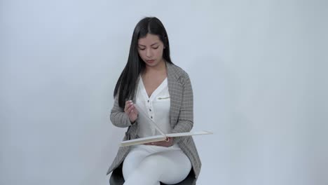Latina-girl-sitting-in-a-chair-reviews-a-book,-turning-each-page-one-by-one,-against-a-white-backdrop,-displaying-concentration-in-her-quiet-study