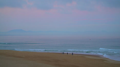 Cinematic-early-morning-pink-purple-dusk-sunrise-surfers-walking-from-a-distance-on-beach-mountain-coastal-landscape-Hoosegor-Seignosse-Biarritz-Basque-Country-Spain-France-WSL-huge-waves-swell-surf