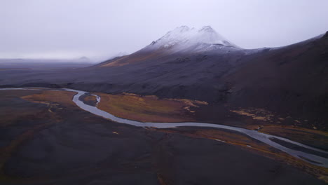 Río-Cruzando-Una-Montaña-Volcánica-Cubierta-De-Nieve-Negra-En-El-Desierto-De-Islandia