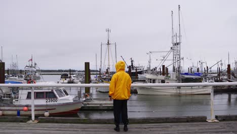 Medium-Shot-of-a-person-in-a-yellow-jacket-walks-dockside-overlooking-a-pier-of-fishing-boats