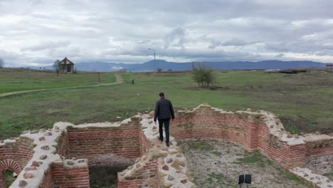 Aerial-view-of-a-man-walking-through-ancient-city-ruins-in-a-large-green-field-on-a-cloudy-day