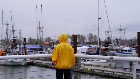 Mittlere-Aufnahme-Einer-Person-In-Einer-Gelben-Jacke,-Die-Am-Hafen-Entlanggeht-Und-Einen-Pier-Mit-Fischerbooten-überblickt