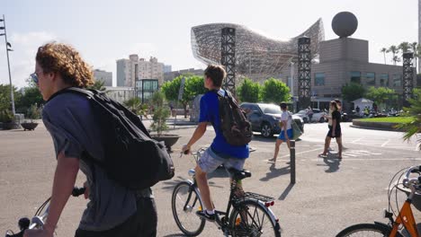 A-group-of-teenagers-ride-bikes-through-the-Port-Olimpic-in-Barcelona,-Spain-with-a-Frank-Gehry-building