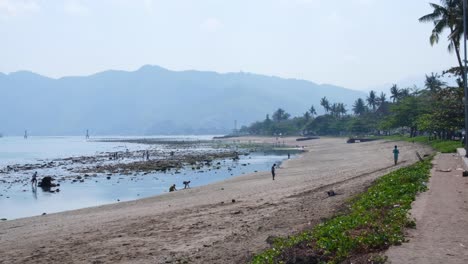 Groups-of-people-on-sandy-beach,-exercising,-playing-and-searching-for-seafood-in-the-shallows-of-ocean-in-capital-city-of-Dili,-Timor-Leste,-Southeast-Asia