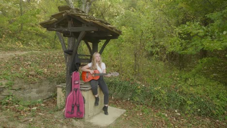 Young-happy-blonde-woman-musician-strums-guitar-woodland-wishing-well