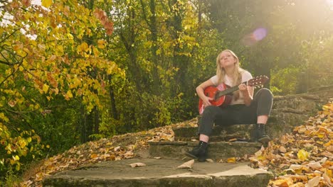 Young-blonde-woman-sits-strums-guitar-autumn-scene-golden-sunshine