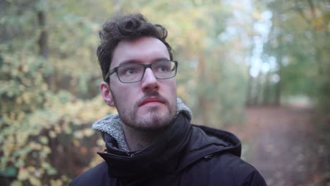 Young-European-man-with-glasses-and-beard-standing-in-a-mixed-autumn-forest,-gazing-into-the-distance