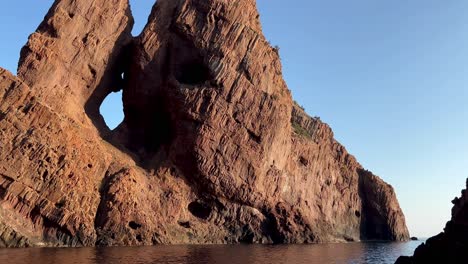 Hole-in-red-rock-of-Scandola-peninsula-nature-reserve-in-summer-season-as-seen-from-moving-boat,-Corsica-island-in-France