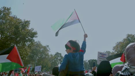 Kleiner-Junge-Bei-Einem-Protest,-Der-Zwischen-Verschiedenen-Schildern-Und-Demonstranten-Eine-Palästinensische-Flagge-Schwenkt