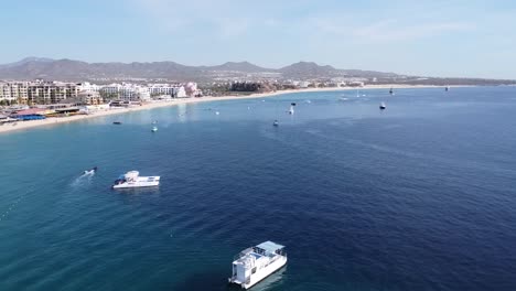 Vista-Aérea-Del-Mar-Azul-Con-Barcos-En-El-Agua,-Instalaciones-Hoteleras-Y-Majestuosas-Montañas-Al-Fondo-En-Un-Día-Soleado-De-Vacaciones-En-La-Playa-Medano-En-Cabo-San-Lucas,-México
