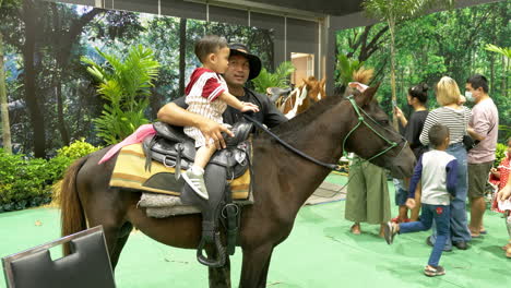 Ein-Kleiner-Junge-Sitzt-Auf-Einem-Pferd,-Während-Der-Pferdeführer-Ihn-Hält,-Sein-Vater-Bereitet-Sich-Darauf-Vor,-Sein-Foto-Zu-Machen,-Im-Hintergrund-Sind-Andere-Familien-In-Einem-Vergnügungspark-In-Bangkok,-Thailand