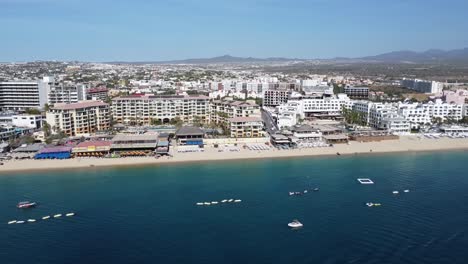 Luftaufnahme-über-Die-Wunderschöne-Küste-Von-Medano-Beach-In-Cabo-San-Lucas,-Mexiko-Mit-Blick-Auf-Das-Blaue-Meer-Mit-Schwimmenden-Booten,-Hotelgebäuden-Und-Einer-Wunderschönen-Strandpromenade-Während-Einer-Tollen-Reise