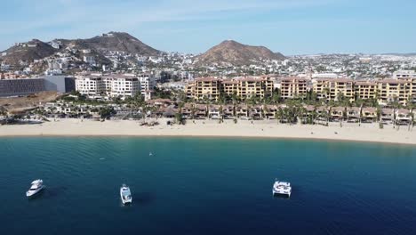 Aerial-shot-over-a-travelers-paradise-over-the-coast-in-front-of-medano-beach-in-cabo-san-lucas,-mexico-with-view-of-the-floating-boats,-gorgeous-beach-and-hotel-complexes-with-mountain-scenery