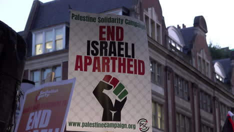 A-Palestine-Solidarity-Campaign-placard-shows-a-first-in-the-red,-green-and-black-flag-colours-and-reads,-“End-Israeli-apartheid”-during-a-protest-at-dusk
