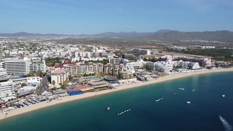 Luftaufnahme-über-Der-Wunderschönen-Küste-Von-Medano-Beach-In-Cabo-San-Lucas-Mit-Blick-Auf-Das-Blaue-Meer-Mit-Schwimmenden-Booten,-Hotelgebäuden-Und-Einer-Wunderschönen-Strandpromenade-Während-Einer-Mexiko-Reise