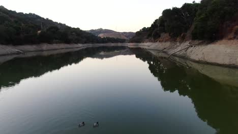 Drone-video-capturing-the-beauty-of-Stevens-Creek-Reservoir-in-Cupertino,-California,-featuring-two-ducks-swimming-on-the-reflective-waters-with-the-mountain-scenery-mirrored-in-the-lake
