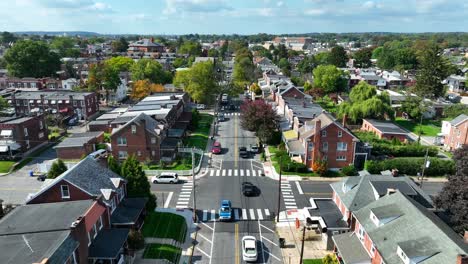 Aerial-view-of-a-suburban-neighborhood-with-brick-houses,-tree-lined-streets,-and-cars-driving-along-a-main-road-on-a-clear-day