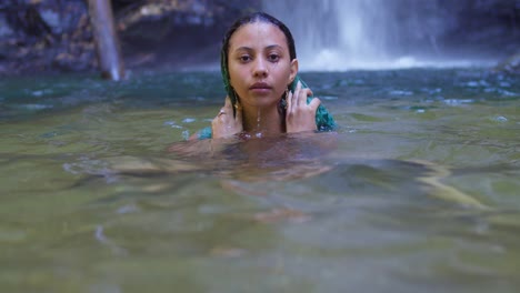 Ein-Mädchen-Mit-Lockigem-Haar-Taucht-Aus-Dem-Wasser-Auf,-Mit-Einem-Fließenden-Wasserfall-Im-Hintergrund