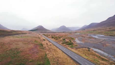 Aerial:-Reveal-of-white-SUV-traveling-along-the-Iceland-Ring-Road-which-is-a-scenic-highway-through-a-picturesque-remote-fjord-area-leading-to-twin-peaks-with-fog-and-haze-in-the-distance
