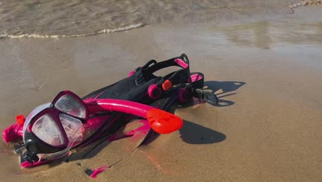 Red-and-fuchsia-fins-mask-and-snorkel-left-on-sandy-beach-with-waves-breaking-on-shore