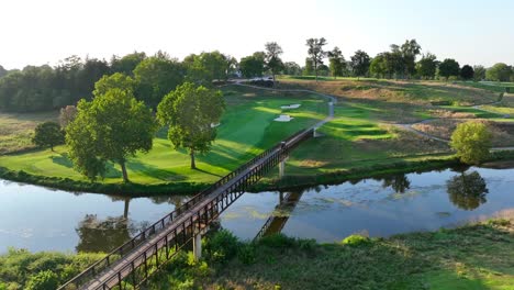 Sunlit-golf-course-in-the-USA,-featuring-a-wooden-bridge-over-a-serene-river,-adjacent-green-fairways,-and-mature-trees