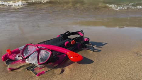 Red-and-black-fins-mask-and-snorkel-left-on-sandy-beach-with-waves-breaking-on-shore