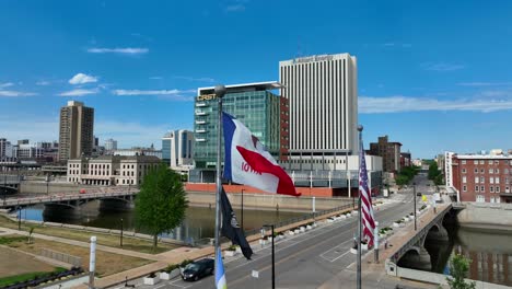 Cedar-Rapids-cityscape-with-a-river-bridge,-towering-buildings-like-Alliant-Energy-and-Crest,-and-fluttering-Iowa-and-US-flags-under-a-blue-sky