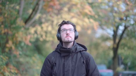 Stylish-European-man-with-a-beard-and-glasses-leisurely-strolls-the-autumnal-suburbs-of-Munich,-deeply-immersed-in-melodies-from-his-headphones,-absorbing-the-seasonal-tranquility