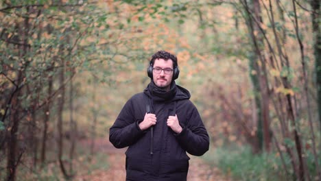 Joyful-bearded-European-man-with-trendy-glasses-stands-amidst-the-hues-of-a-mixed-autumn-forest,-radiating-happiness-and-warmth-upon-seeing-viewers,-emanating-pure-positive-vibes