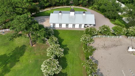Experience-the-epitome-of-refined-country-living-with-this-stunning-aerial-view-of-a-pristine-white-barn-nestled-within-an-exclusive-club,-framed-by-vibrant-Crepe-Myrtles-in-full-bloom