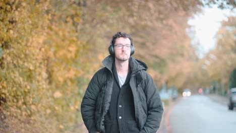 Stylish-European-man-with-a-beard-and-glasses-leisurely-strolls-the-autumnal-suburbs-of-Munich,-deeply-immersed-in-melodies-from-his-headphones,-absorbing-the-seasonal-tranquility